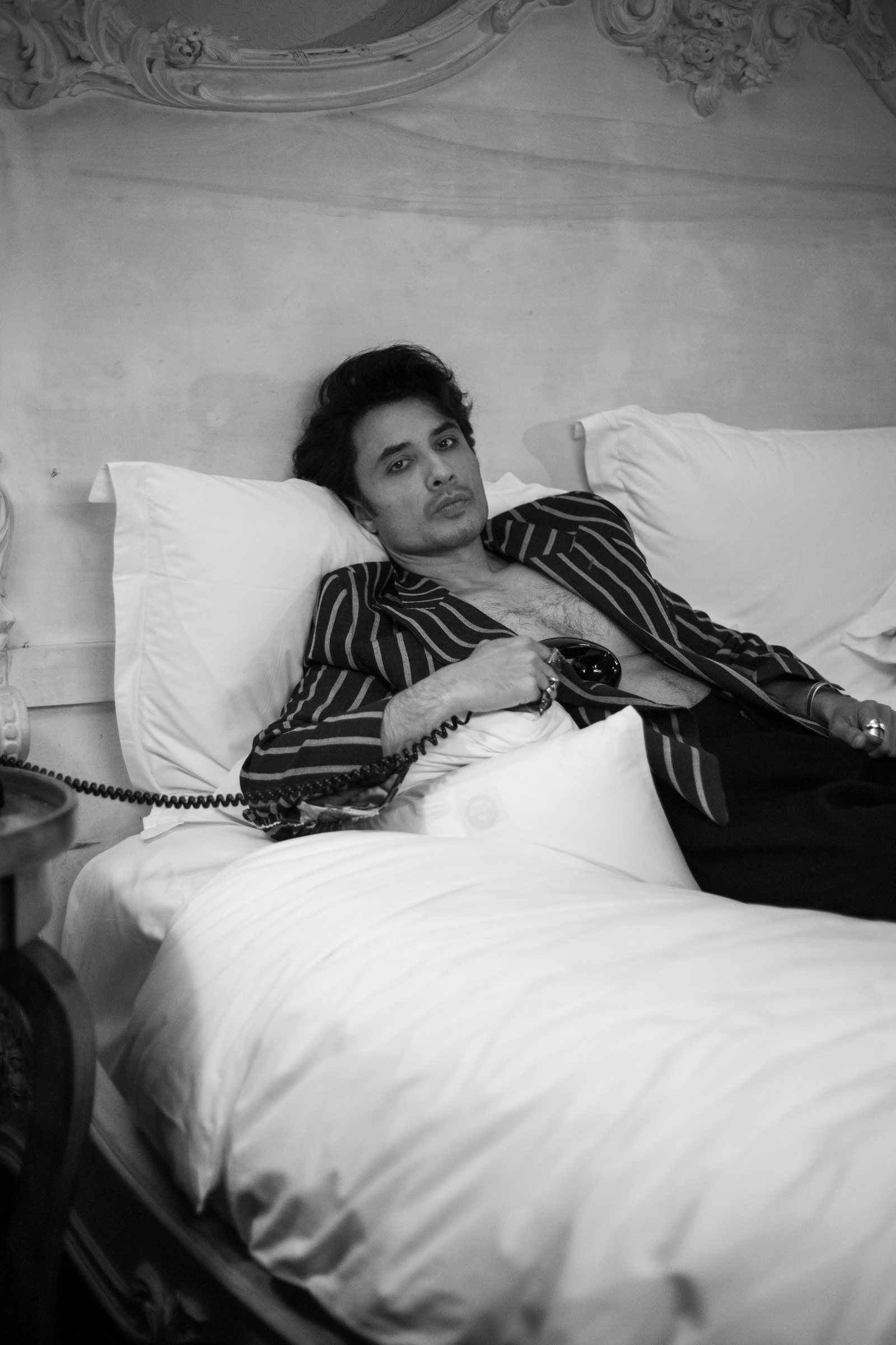 Ali Zafar lying back on a hotel bed in a striped jacket, a telephone receiver in his hand, looking into the camera
